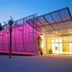 Manarat Al Saadiyat entrance at dusk, featuring a modern building with a pink perforated facade, a patterned metal canopy over the glass doors, and palm trees in the foreground.